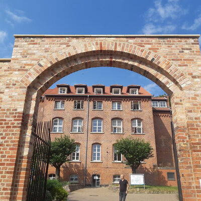 Ein rotes Backsteingebäude mit blauen Himmel ist durch einen gemauerten Torbogen zu sehen, während eine Person vorbeigeht.