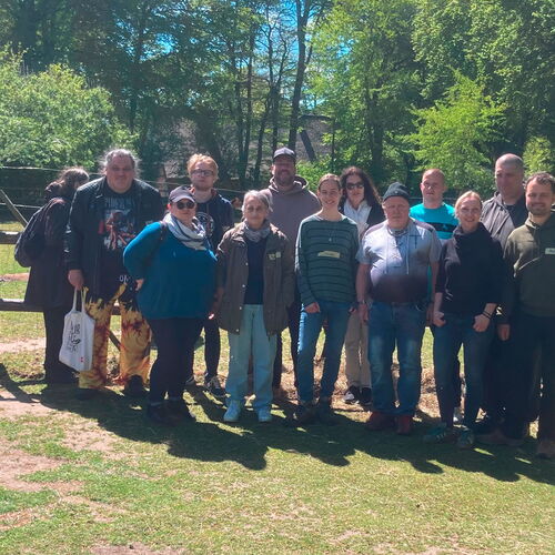 Eine Gruppe von Menschen posiert fröhlich auf einer Wiese, während ein Alpaka im Hintergrund steht.  | © Name