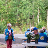 Eine Frau in blauer Weste geht an einem Tisch mit Leuten vorbei, die im Freien bei einem Treffen sitzen.