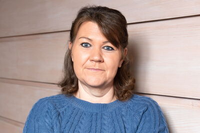 Eine Diakonie-Mitarbeiterin in einem blauen Pullover lächelt vor einer hellen Holzwand.