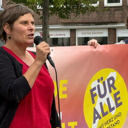 Eine Frau hält eine Rede auf einer Veranstaltung mit einem Banner, das "FÜR ALLE" zeigt.