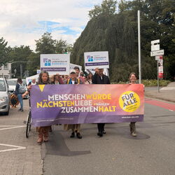 Menschen marchieren mit einem Banner für Nächstenliebe und Zusammenhalt auf einer Straße bei einer Demonstration.