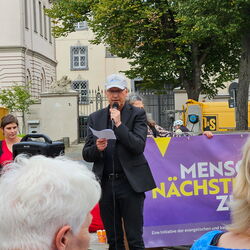 Eine Person spricht bei einer Veranstaltung für "Menschen nächst" vor einem Publikum mit sichtbarem Banner.