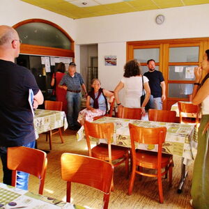 Eine Gruppe von Menschen steht in einem Raum mit Tischen und kommuniziert lebhaft miteinander. | © Name