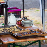 Auf einem Holztisch stehen Kuchenbleche, Geschirr und ein Wasserkocher für eine Buffetvorbereitung im Freien.