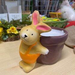 Ein fröhlicher Hase mit rosa Ohren und orangefarbener Weste steht neben einem braunen Blumentopf auf einem Tisch.
