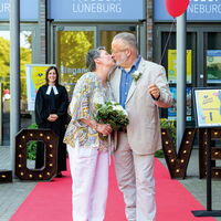 Regina und Marc küssen sich auf dem roten Teppich vor dem Museum Lüneburg, während die Vikarin lächelt.
