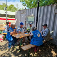 Menschen sitzen bei sonnigem Wetter an Holztischen vor einem Container, während ein Zug im Hintergrund vorbeifährt.