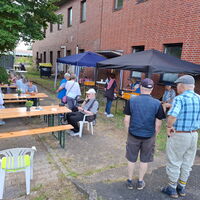 Menschen sitzen an Holztischen und unterhalten sich bei einem geselligen Treffen im Freien vor einem Backsteingebäude.