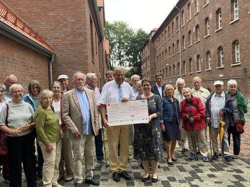 Eine Gruppe von Senioren steht zusammen mit einem Scheck in einer historische Gasse. | © Stiftung Dimm