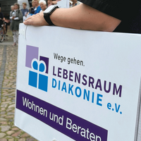 Demonstration für Wohnraum und Beratung, mit Menschen und einem Schild der Lebensraum Diakonie e.V. in der Öffentlichkeit.