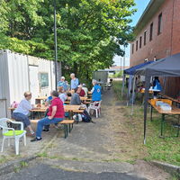 Menschen sitzen an Holzbänken unter einem Pavillon im Freien und genießen ein geselliges Beisammensein an einem sonnigen Tag.