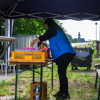 Eine Person in blauer Weste bereitet Getränke auf einem Tisch in einem Garten unter einem Zelt vor.