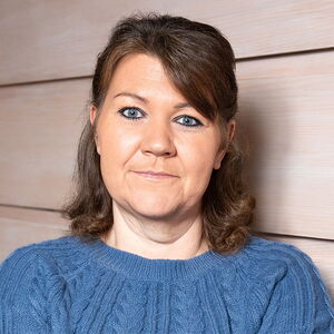 Eine Frau mit blauer Strickjacke blickt freundlich in die Kamera vor einer hellen Wand.