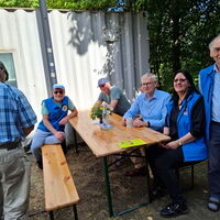 Eine Gruppe von Menschen sitzt entspannt an einem Holztisch im Freien, umgeben von Bäumen.