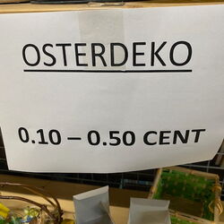 Ein Schild mit der Aufschrift „Osterdeko“ und Preisen zwischen 0,10 und 0,50 Cent ist auf einem Tisch sichtbar.