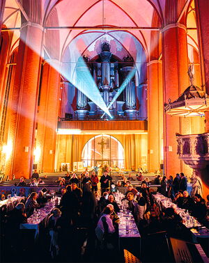 Menschen sitzen in einer festlich beleuchteten Kirche, während Lichtstrahlen auf eine große Orgel fallen.