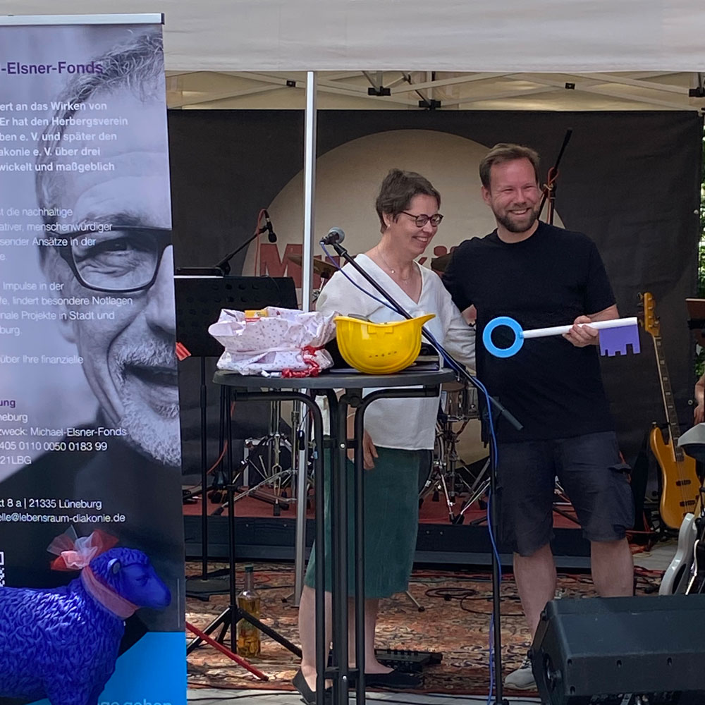 Zwei Personen feiern die Einweihung mit einem großen Schlüssel und einem Gelbes Helm beim Event des Michael-Elsner-Fonds.