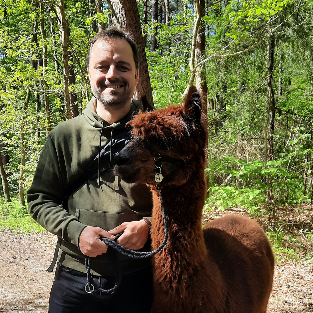 Ein Mann steht im Wald und hält ein braunes Alpaka an der Leine, beide lächeln in die Kamera. | © Name