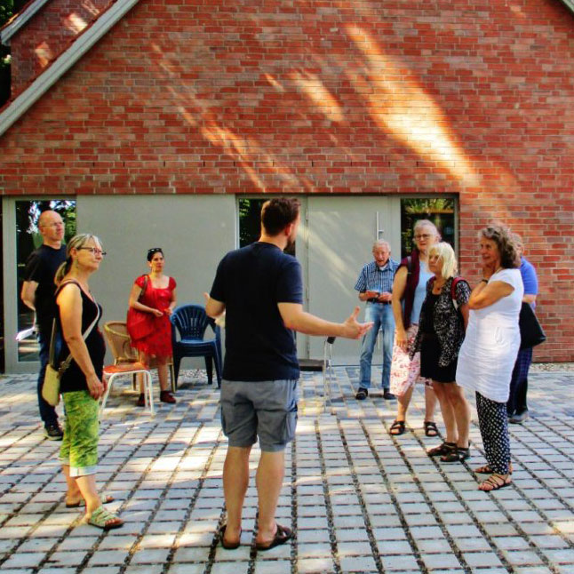 Eine Gruppe von Menschen hört einem Redner vor einer Backsteinwand zu, während Sonnenstrahlen auf den Platz fallen.