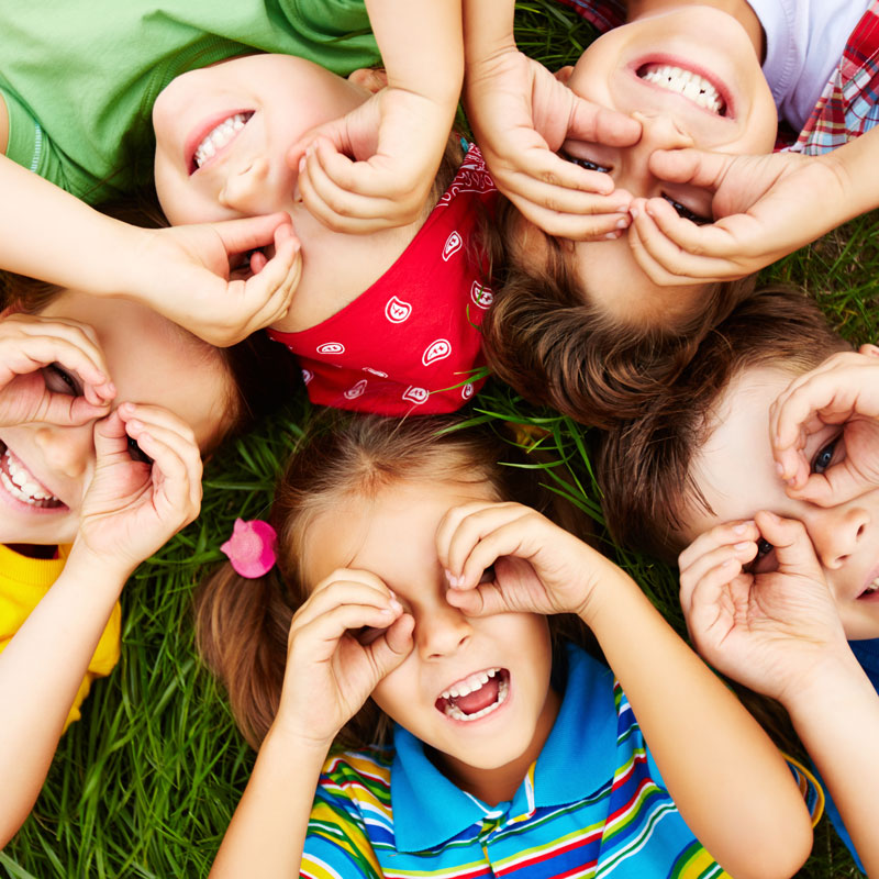 Sechs Kinder liegen im Gras und formen mit ihren Händen lustige Gesichter, während sie lachen und spielen.