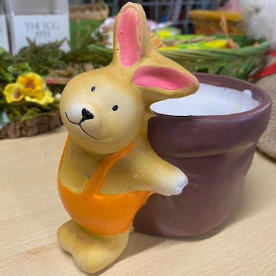 Ein fröhlicher, gelber Hase mit rosa Ohren steht an einem Blumentopf und verbreitet Osterstimmung.