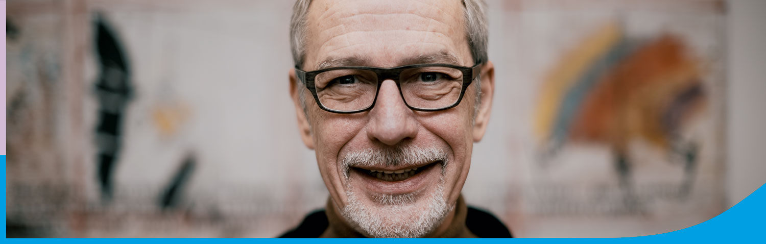 Lächelnder Mann mit Brille vor einer bunten Wandgestaltung, die eine kreative Atmosphäre vermittelt.