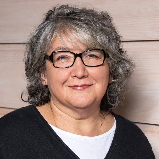 Eine lächelnde Frau mit grauen Locken und Brille posiert vor einer hellen Holzoberfläche.