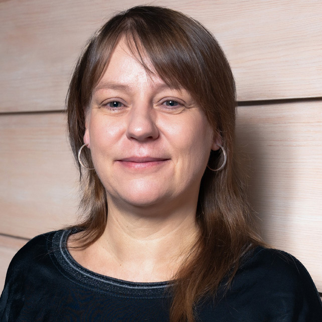 Eine Frau mit lässiger Frisur und Ohrringen lächelt vor einer Holzpaneel-Wand.