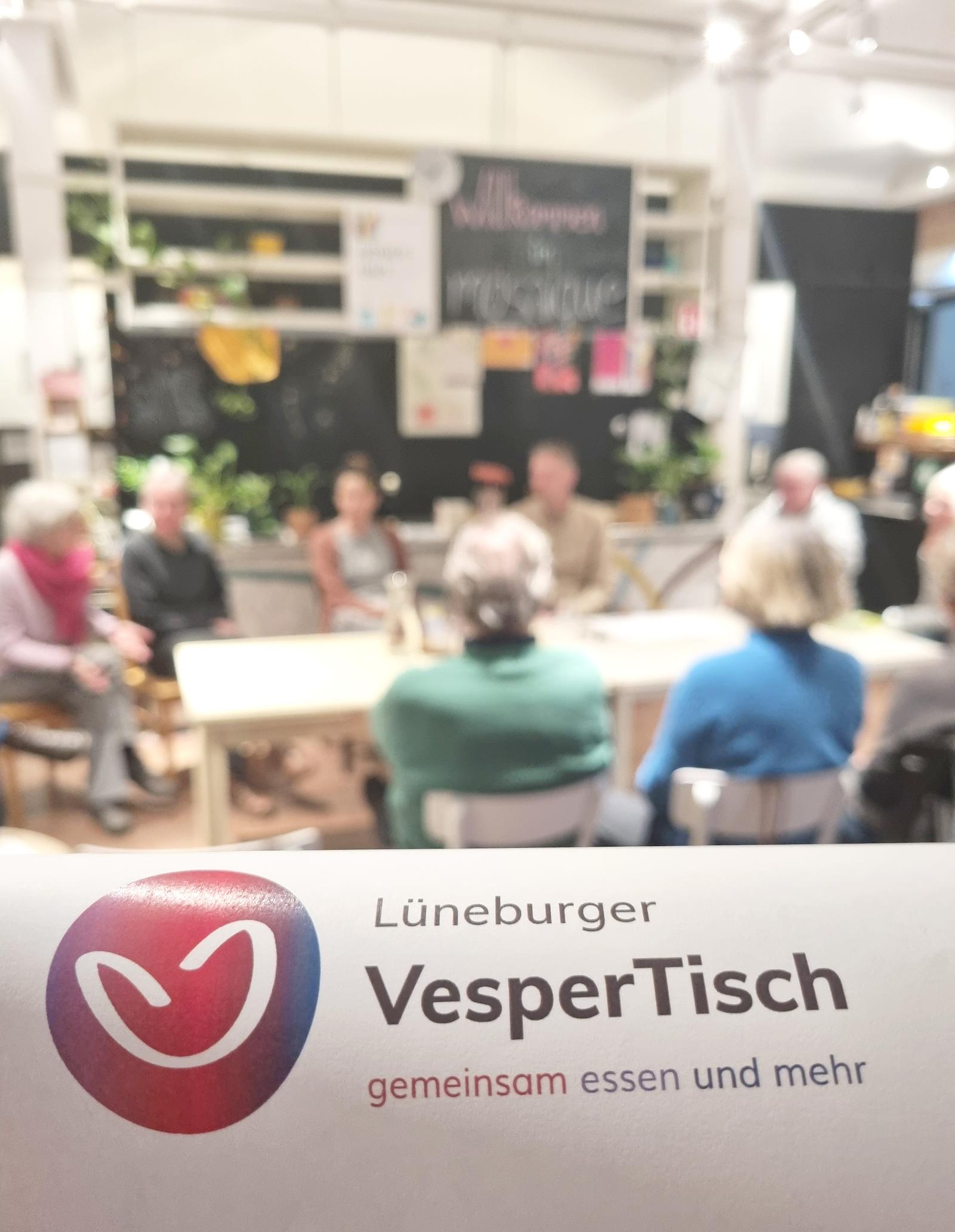 Eine Gruppe von Menschen sitzt um einen Tisch und diskutiert beim Lüneburger VesperTisch über gemeinsames Essen und mehr.